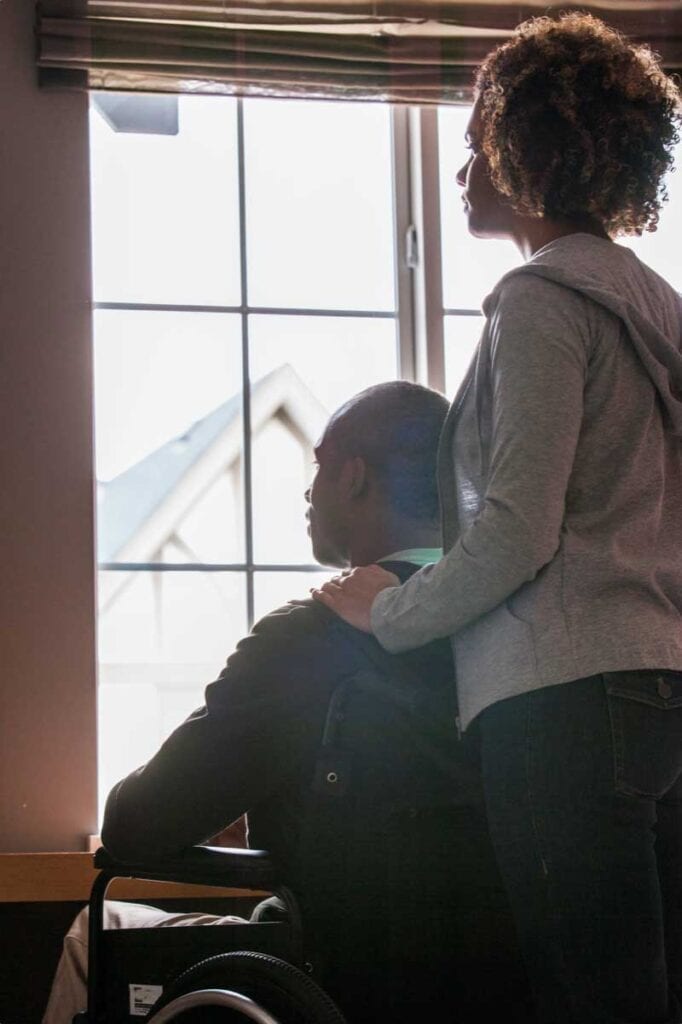 a woman standing and man in a wheelchair look out of window