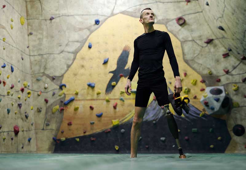 veteran with prosthetic left leg stands in front of rock wall, looks off into distance